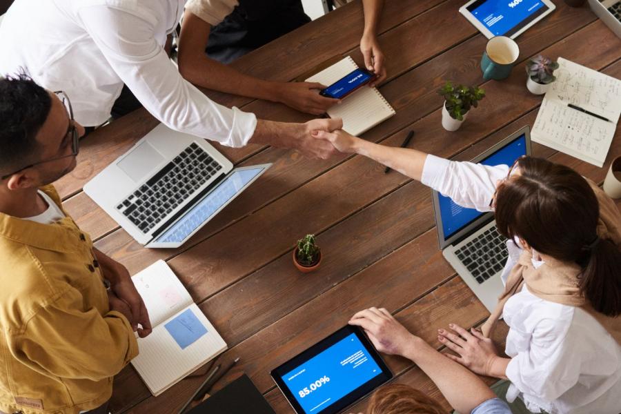  In una ripresa dall'alto, due persone si stringono la mano a un tavolo di legno scuro, circondate da laptop, tablet e quaderni. Altre mani sono visibili intorno al tavolo, suggerendo la presenza di più individui che partecipano a una riunione o una collaborazione.