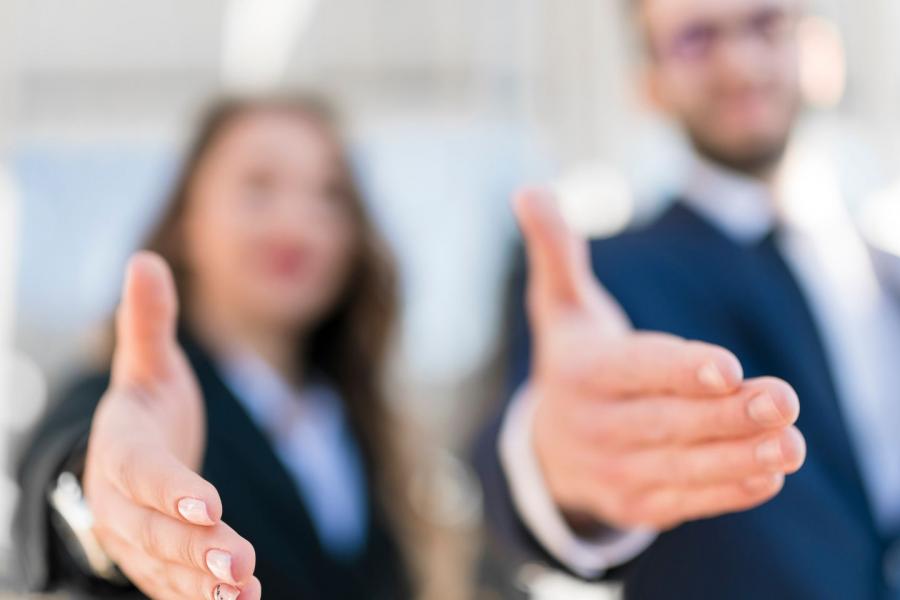 Un uomo e una donna in abiti formali protendono la mano verso l’osservatore in un gesto di accoglienza, saluto o offerta di aiuto. I volti sono sfocati, mentre le mani in primo piano sono a fuoco, enfatizzando il gesto